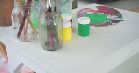 Bright acrylic paint jars and brush-filled mason jars on white studio table closeup