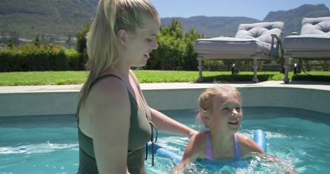 Mother supporting daughter learning to swim in backyard pool with blue noodle on sunny summer day