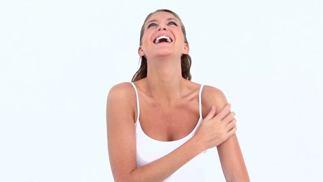 Woman enjoying her skincare routine, moisturizing her arm with joy and laughter against a minimalist white background. Perfect for wellness, beauty, and self-care advertisements or blog features exploring happiness and skincare routines.