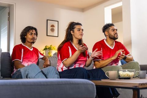 Friends cheering on sofa in matching red jerseys watching live sports match at home