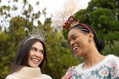 Diverse friends wearing merry christmas headbands laughing in garden in cozy knit sweaters