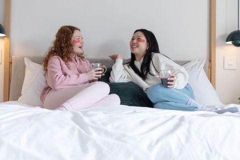 Diverse female friends relaxing on bed wearing under-eye patches and sipping tea