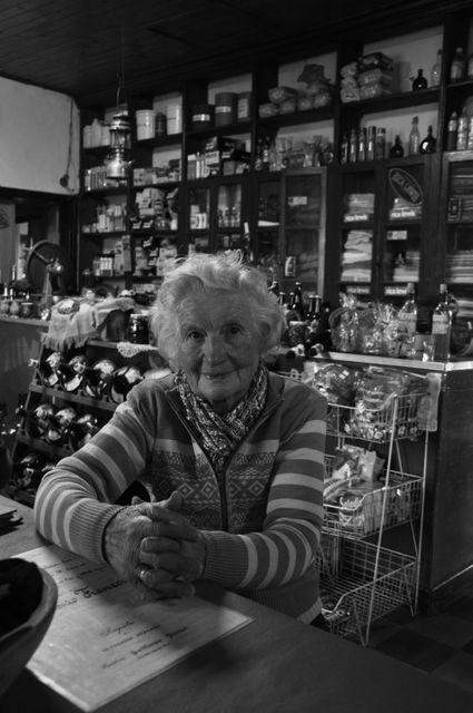Elderly woman sitting behind counter in traditional grocery store with shelves filled with various merchandise in background, captured in black and white. Ideal for use in articles or advertisements focusing on nostalgia, heritage small businesses, community stories, or historical records. Could also be used in promotions for retro-themed events or local heritage tourism.