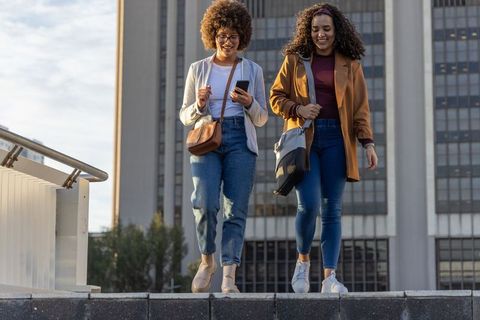 Two african american women walking down steps in city plaza using smartphone and carrying bags