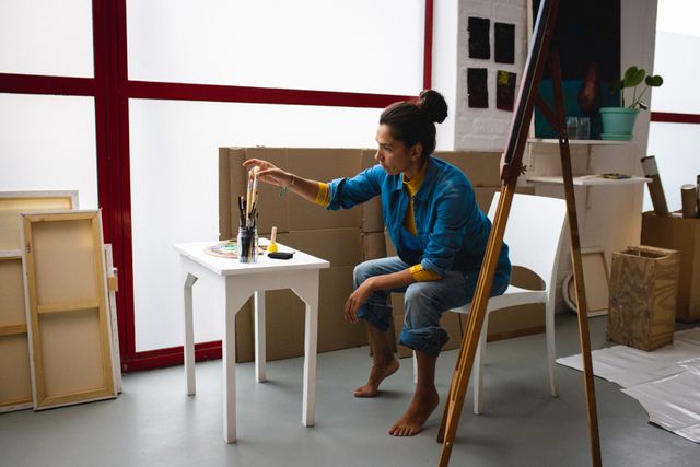 Biracial female artist with painting equipment sitting on chair in creative office. Female artist studio and painting concept.