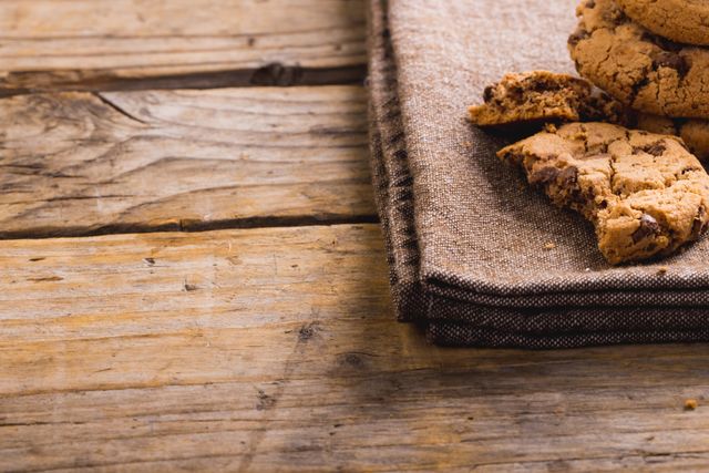 This image shows a close-up of chocolate chip cookies placed on a piece of burlap on a wooden table. The rustic setting and the texture of the burlap and wood create a warm, homemade feel. Ideal for use in food blogs, recipe websites, bakery promotions, or advertisements for homemade goods. The copy space allows for easy addition of text or logos.