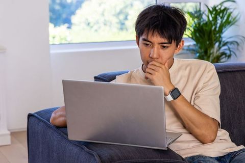 Asian man working from home on laptop, contemplative on sofa near window plants