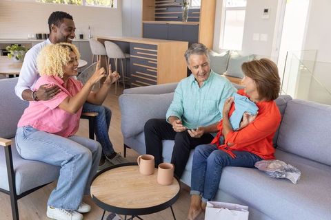 Multigenerational diverse family welcoming newborn at home, senior cradling baby on sofa