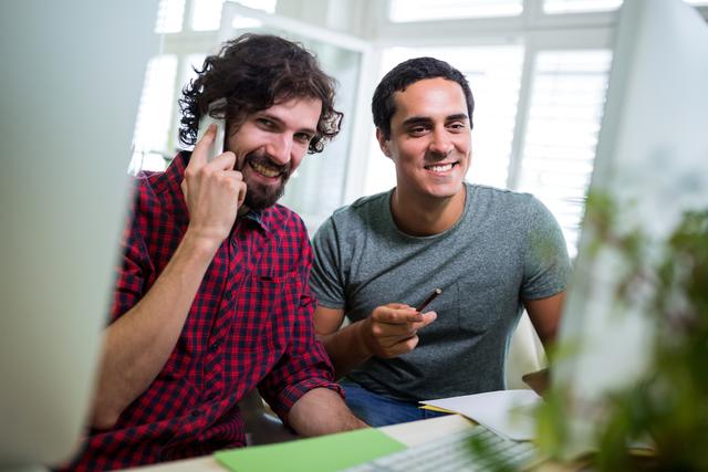 Male graphic designer talking on mobile phone while coworker looking at computer in office