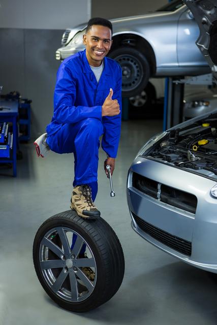 Mechanic in blue coveralls smiling and giving thumbs up while standing on a tire in an auto repair garage. Ideal for illustrating automotive services, car maintenance, and professional repairman themes. Suitable for use in advertisements, brochures, and websites related to auto repair shops and mechanical services.