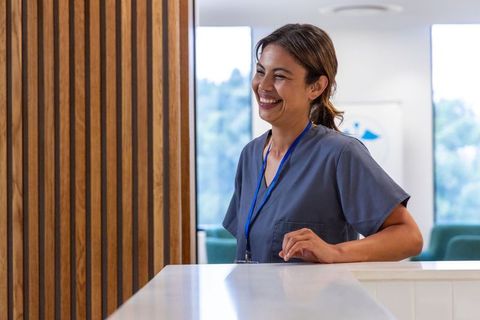 Smiling healthcare receptionist wearing scrubs and lanyard at modern clinic reception desk