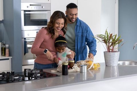 Multicultural family hugging daughter at modern kitchen island unpacking groceries and jars