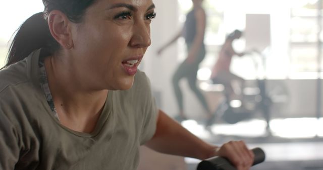 A woman shows determination and strength while exercising on a rowing machine at the gym, with blurred figures in the background exercising as well. This can be used for highlighting dedication, promoting gym memberships, fitness programs, or healthy lifestyle campaigns.