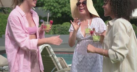 Diverse women laughing and sipping strawberry cocktails by poolside wearing sunhat and sunglasses