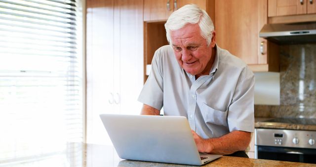 An elderly man interacts with his laptop in a contemporary home kitchen setting, showcasing the blend of technology and daily living for seniors. Ideal for websites focusing on senior care, technology adoption among the elderly, home lifestyles, work-from-home culture among retirees, and independent living for older adults. Can be used to illustrate articles on aging, retirement activities, and remote work environments.