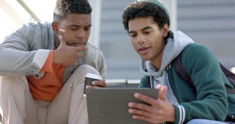 African american college friends sharing tablet on campus steps, studying with coffee