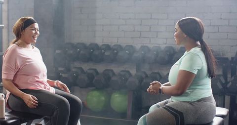 Diverse women chatting on gym benches wearing activewear and smiling during rest