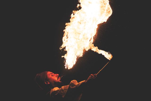Dramatic scene of a fire performer breathing flames into dark night sky. Ideal for use in topics related to stunt performances, pyrotechnics, street entertainment, and dangerous acts. Suitable for promoting events, festivals, or showcasing the excitement of performers.