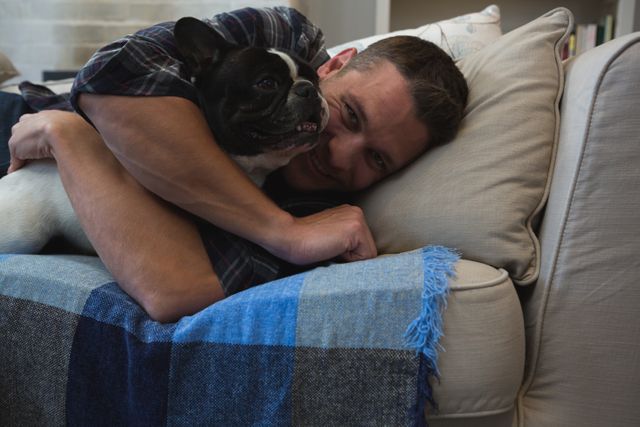 Man enjoying quality time with his French Bulldog on a comfortable sofa in a cozy living room. Ideal for use in articles or advertisements about pet care, home life, relaxation, and companionship. Perfect for illustrating themes of love, bonding, and the joy of having pets.