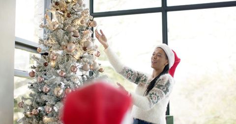 Woman in santa hat decorating flocked christmas tree with rose gold ornaments by window