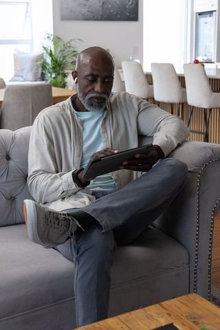 Mature african american man relaxing on sofa using tablet wearing earbuds in contemporary living roo