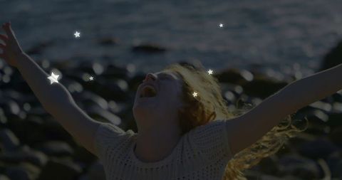 Laughing woman raising arms on pebble beach with sunlit hair and star overlays