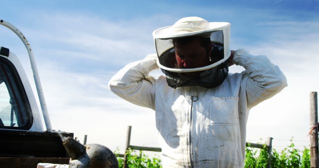 Beekeeper in protective suit preparing for work with beehives. Rural vineyard in background offers serene environment for beekeeping. Suitable for articles on agriculture, beekeeping practices, rural lifestyles, honey production, and outdoor occupations.
