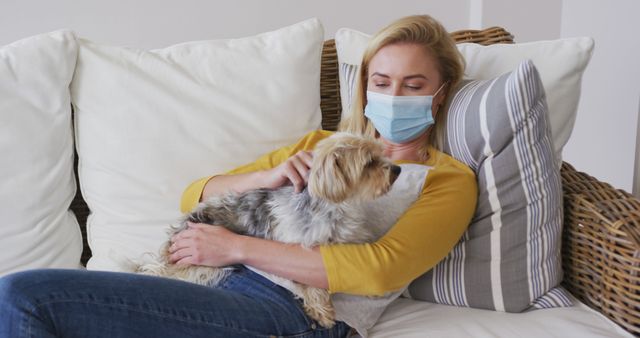 Woman wearing a medical mask relaxing on a white couch with comfortable pillows while holding a small dog. Ideal for themes around COVID-19 precautions, indoor comfort, pet companionship, and home safety measures. Suitable for use in articles or promotions related to health, home lifestyle, and pet care.