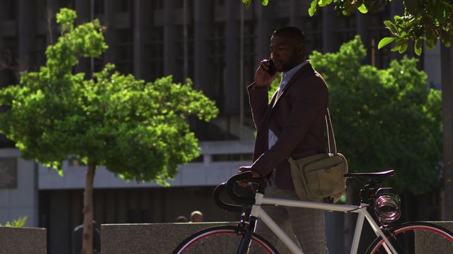 African American man holding bicycle and talking on smartphone in urban setting with green trees in background. Ideal for depicting concepts like urban commuting, technology on the go, business communication, and modern digital nomad lifestyle.