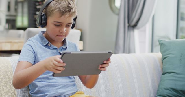 Boy wearing headphones sitting on a sofa using a tablet. Useful for educational, technology, and family lifestyle campaigns as well as depictions of modern childhood activities in home settings.