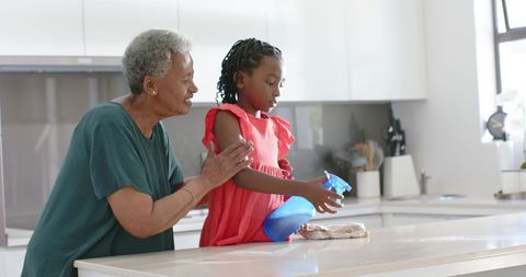 Grandmother guiding granddaughter cleaning kitchen counter, intergenerational caregiving