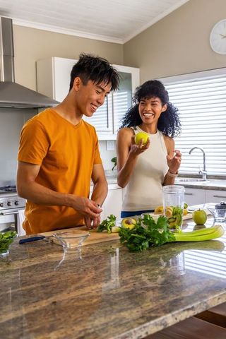 Multiethnic couple making green smoothie in bright kitchen chopping apples and celery