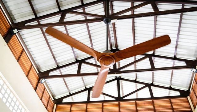 Wooden three-blade ceiling fan in high-roof pavilion featuring exposed trusses and louvers