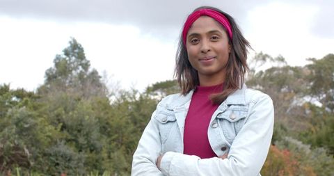 Confident Indian woman standing wearing magenta top and denim jacket with headband in park
