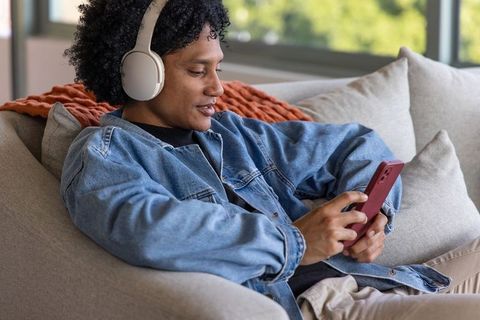 African american man relaxing on sofa wearing headphones and denim jacket using smartphone