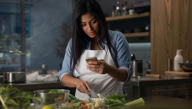 Home cook chopping vegetables while using smartphone in modern stainless-steel kitchen