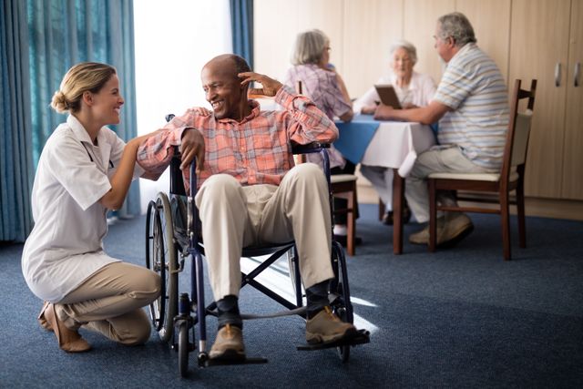 Smiling female doctor kneeling by disabled senior man sitting on wheelchair at retirement home