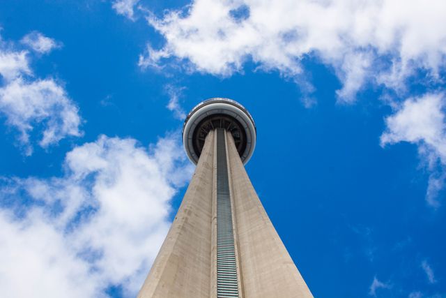 This photo depicts the CN Tower from a low angle, emphasizing its height against a blue sky with fluffy clouds. The perspective highlights the architectural marvel of this urban landmark in Toronto. Ideal for use in travel brochures, architectural presentations, website headers, and articles relating to city attractions or architectural feats.