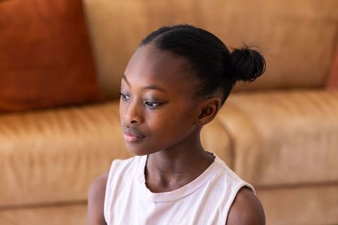 Thoughtful young black girl sitting on beige sofa looking away in warm home portrait