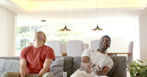 Friends playing video games on sofa laughing in modern bright open-plan living room