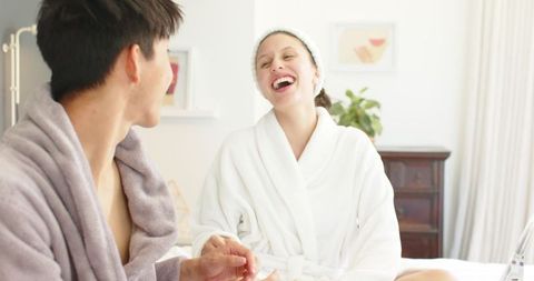 Asian man and african american woman laughing during morning spa manicure, bright bedroom