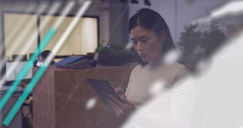 Asian woman tapping tablet in modern open-plan office with glass reflections, focused