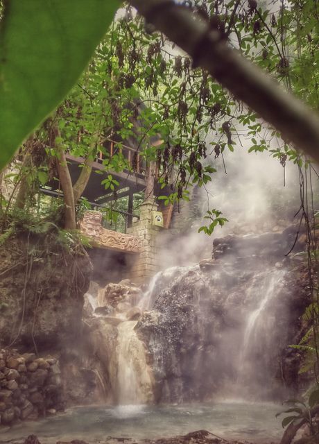 Misty waterfall cascading through lush tropical jungle with rustic wooden structure in background. Ideal for travel brochures, nature conservation campaigns, wellness retreat advertising, relaxing ambiance imagery.