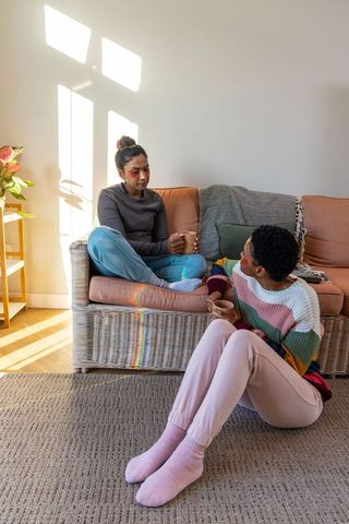 Two diverse women relaxing and chatting in cozy sunlit living room while holding warm ceramic mugs