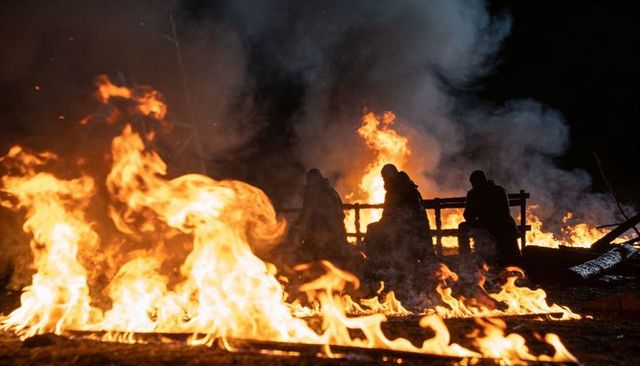 Silhouetted figures crouching behind wooden barrier amid roaring fire at night