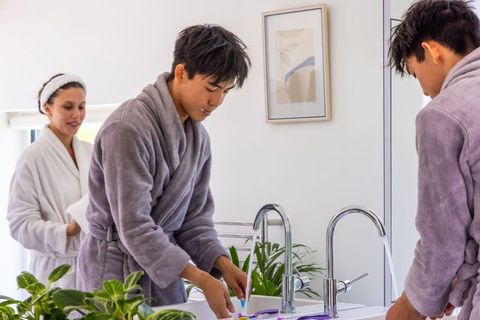 Couple sharing morning routine, asian man brushing teeth at vanity while partner holds towel