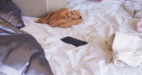 Black smartphone lying on rumpled white bedding with casual clothes and soft natural light