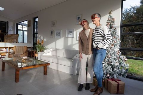 Two women celebrating christmas wearing festive novelty glasses by decorated tree smiling
