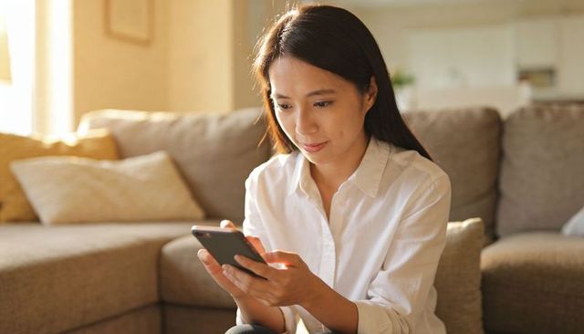 Woman scrolling smartphone on beige sofa in sunlit cozy living room