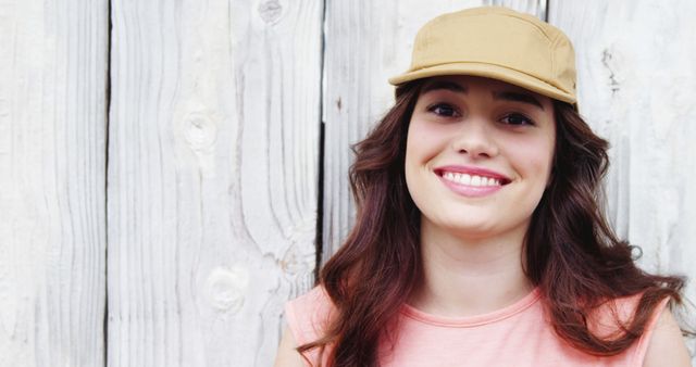 Woman smiling while wearing a beige cap and casual clothes in front of a wooden background. Ideal for lifestyle blogs, outdoor fashion advertisements, casual wear promotions, and summer activity websites.
